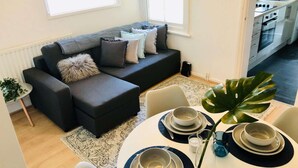 Un salon moderne avec un canapé d’angle gris, une table à manger dressée avec des assiettes et des bols, et une grande plante verte.