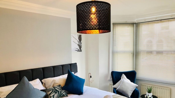 Une chambre d’hôtel avec un grand lit, plusieurs oreillers, une table de chevet avec une lampe et une fenêtre avec des stores.