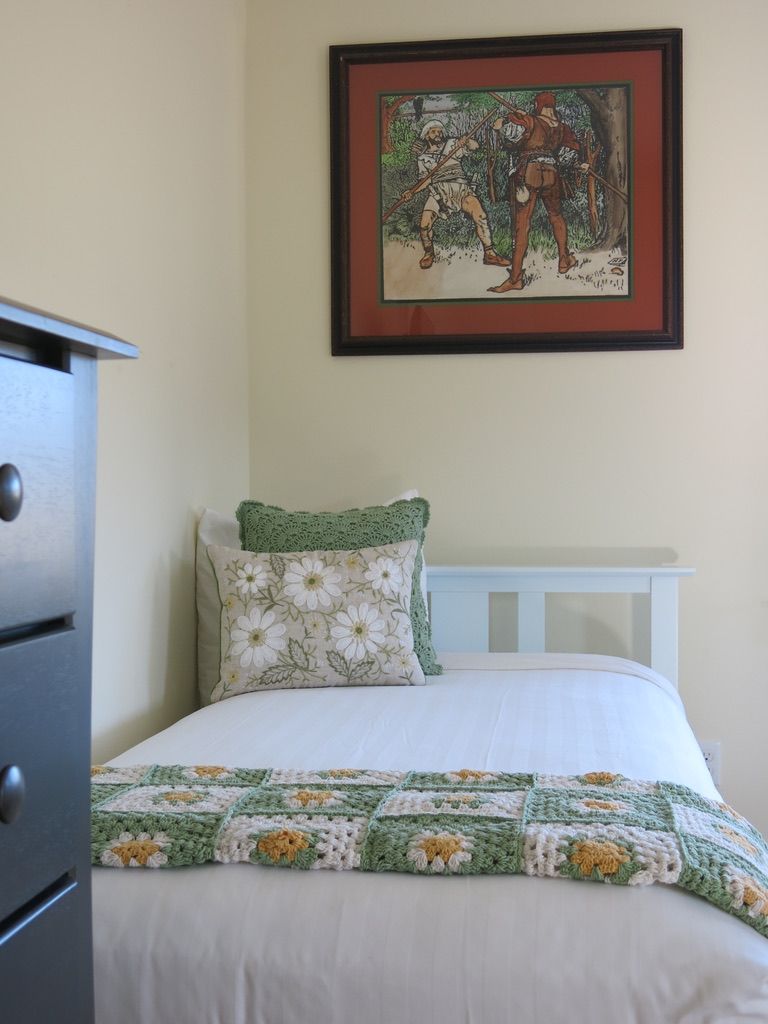 A bedroom with a bed, a framed picture on the wall, a dresser, and a green crocheted blanket.