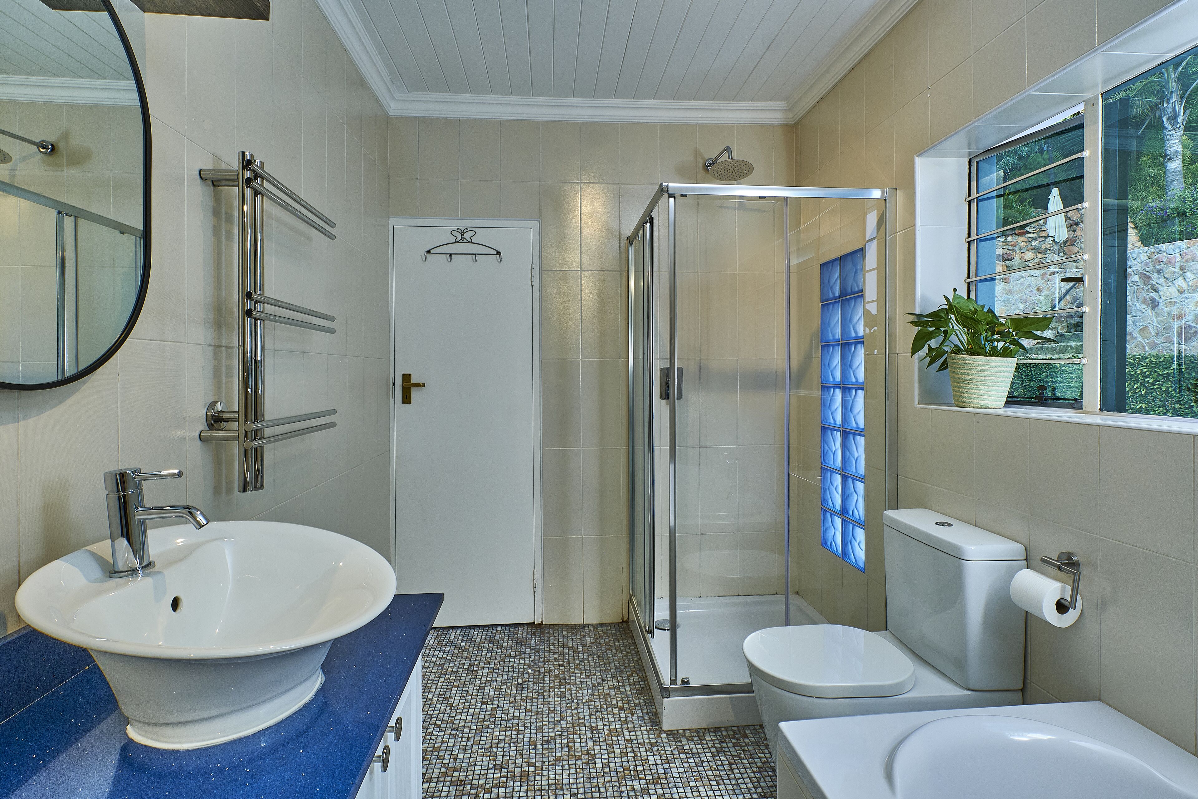 A bathroom with a white sink, a glass shower enclosure, a toilet, and a window with a view of greenery.