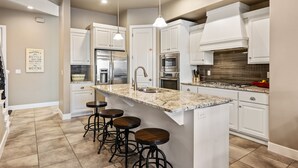 A modern kitchen with white cabinets, a granite countertop, a stainless steel refrigerator, and a built-in oven. There are three bar stools and pendant lights above the island. - Zion's Edge: Two Pools and Hot Tubs, Clubhouse Gym (Washington)