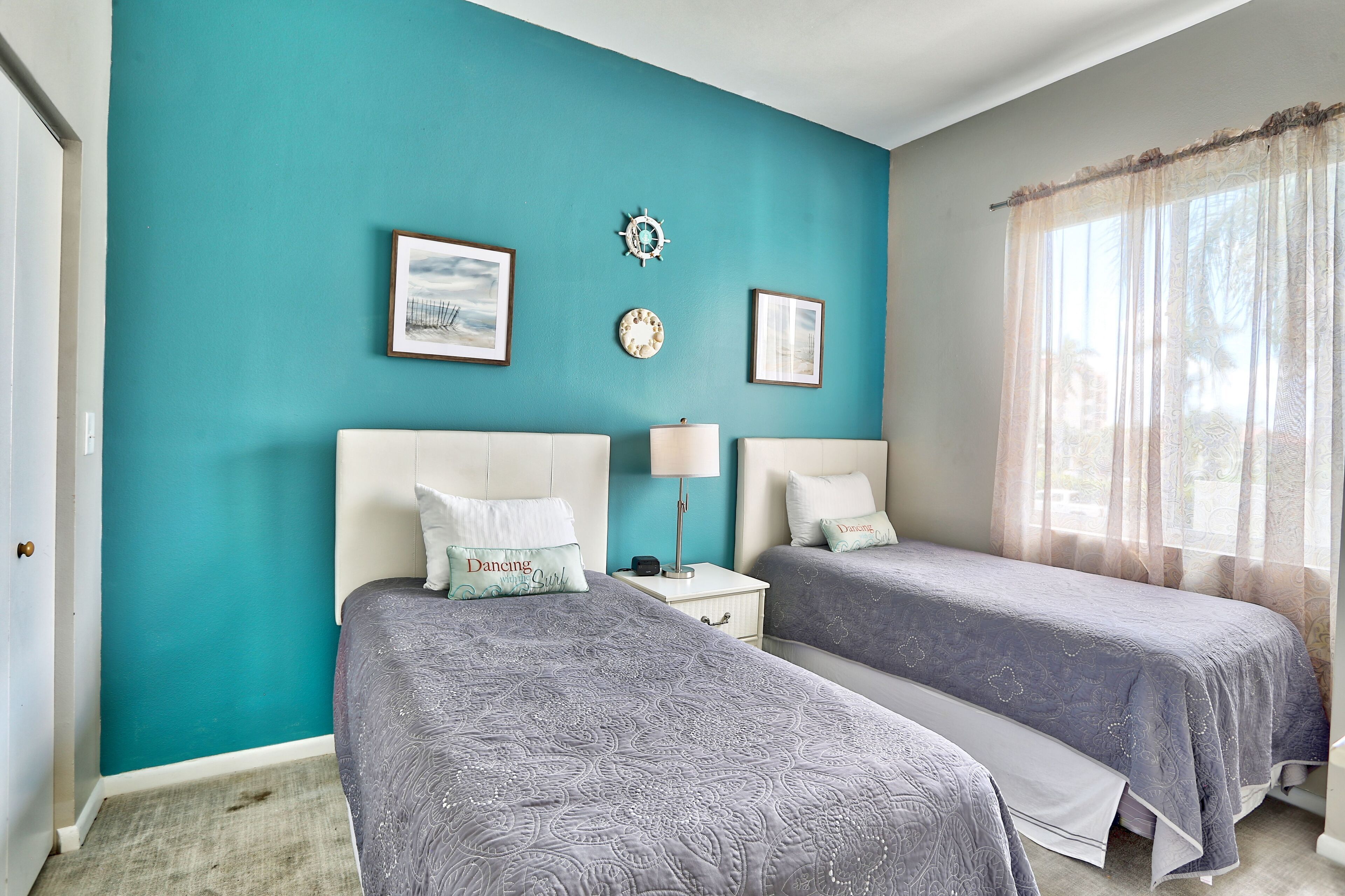 A bedroom with two beds, teal walls, framed pictures, a clock, and a window with sheer curtains.