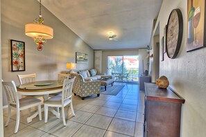 A spacious living area with a dining table, chairs, a sofa, and a coffee table. There is a chandelier, a ceiling fan, and artwork on the walls.