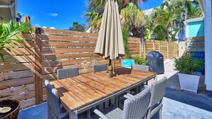 An outdoor patio with a wooden table and wicker chairs, surrounded by a wooden fence and palm trees.