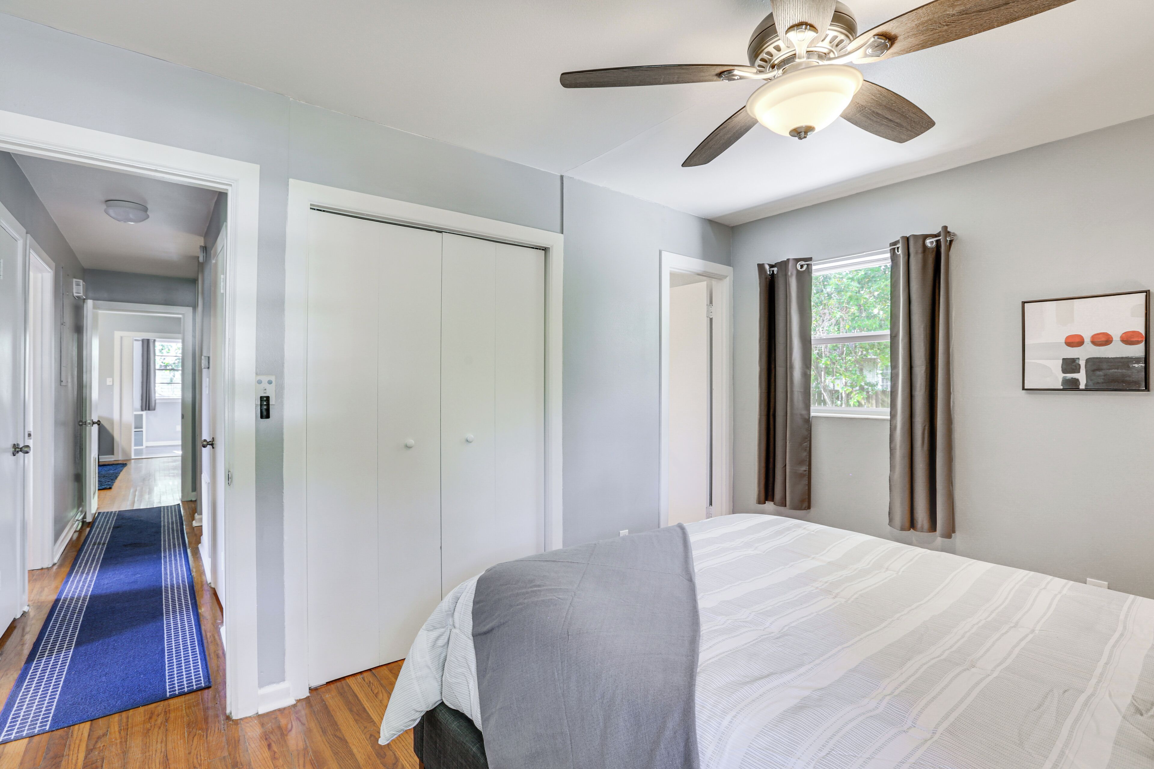 Un dormitorio con una cama, un ventilador de techo, una ventana con cortinas y un pasillo que conduce a otra habitación.