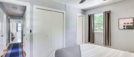Un dormitorio con una cama, un ventilador de techo, una ventana con cortinas y un pasillo que conduce a otra habitación.