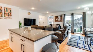 A modern living room with a central island, a flat-screen TV, a sofa, and a dining area with a chandelier. - Modern Elizabeth Apartment ~ 1 Mi to Train Station (Elizabeth)