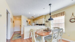 A dining area with a wooden table and white chairs, a kitchen in the background, and a window with blinds.