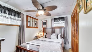 Ein Schlafzimmer mit Bett, Deckenventilator, Nachttisch, Spiegel und Fenster mit Vorhängen.