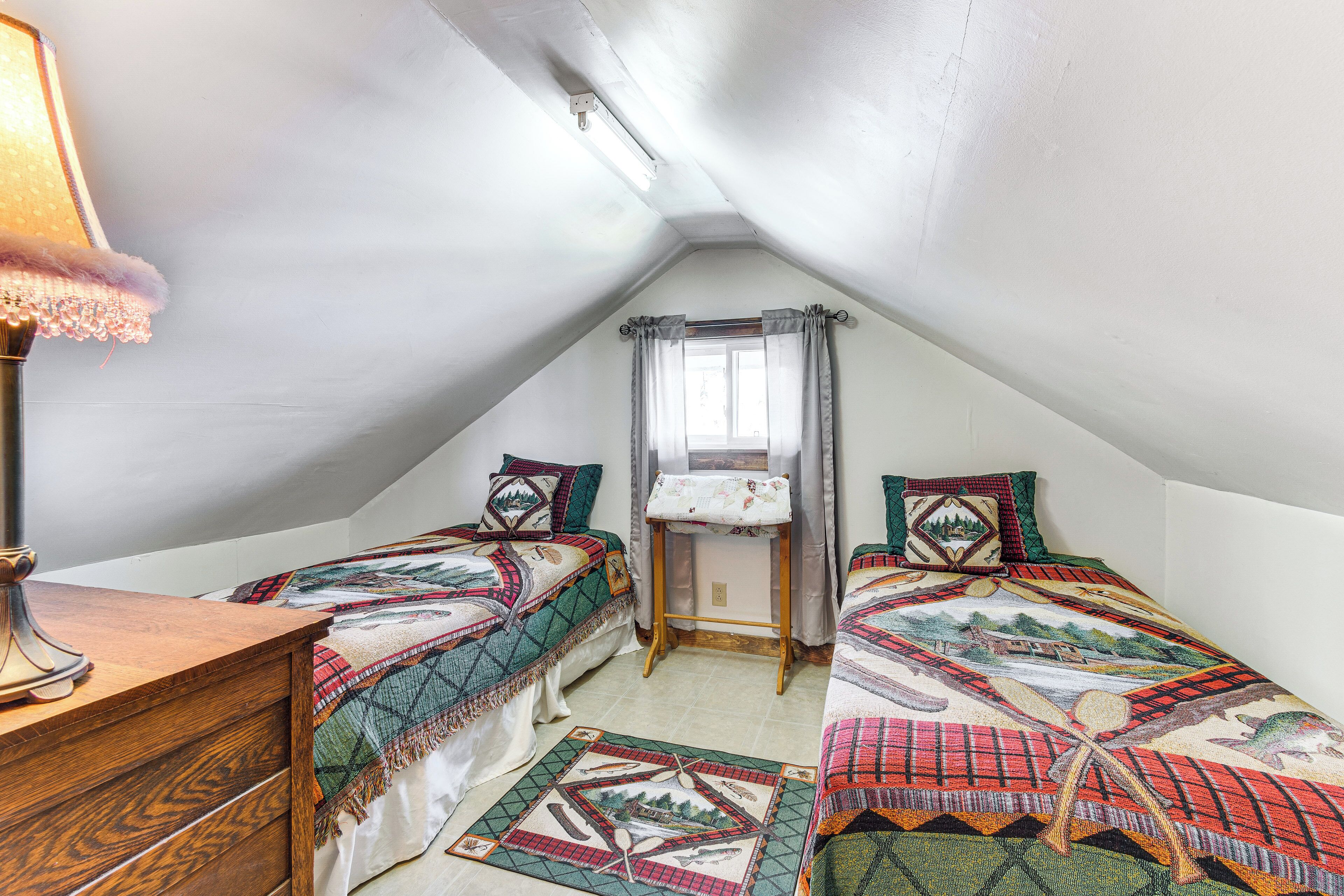 A small, cozy attic bedroom with two single beds, a wooden dresser, a desk, and a window with curtains.