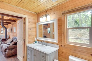 L'intérieur d'un chalet en bois est aménagé avec un fauteuil inclinable en cuir, une salle de bain dotée d'un grand miroir et une fenêtre donnant sur une zone boisée.