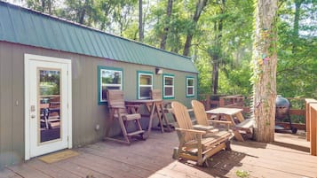 Une terrasse en bois avec des chaises Adirondack et des tables de pique-nique, un barbecue en métal et un bâtiment doté d’un toit végétalisé et de fenêtres.