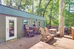 Une terrasse en bois avec des chaises Adirondack et des tables de pique-nique, un barbecue en métal et un bâtiment doté d’un toit végétalisé et de fenêtres.