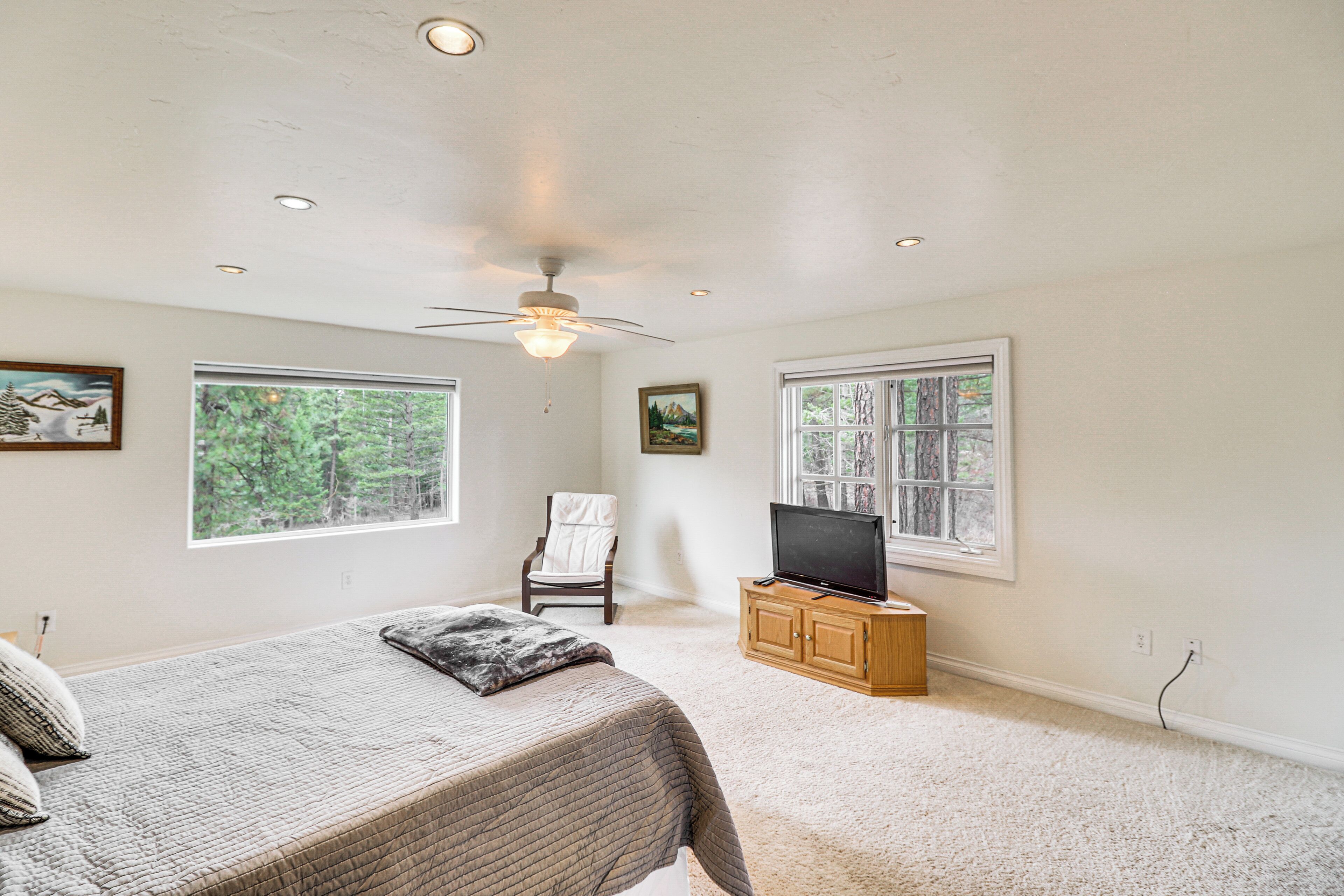A bedroom with a bed, a chair, a television on a wooden cabinet, and large windows with a view of trees.