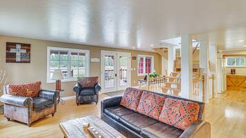 A spacious living room with a large leather sofa, two armchairs, and a wooden coffee table. There are multiple windows allowing natural light, and a staircase leading to an upper level.