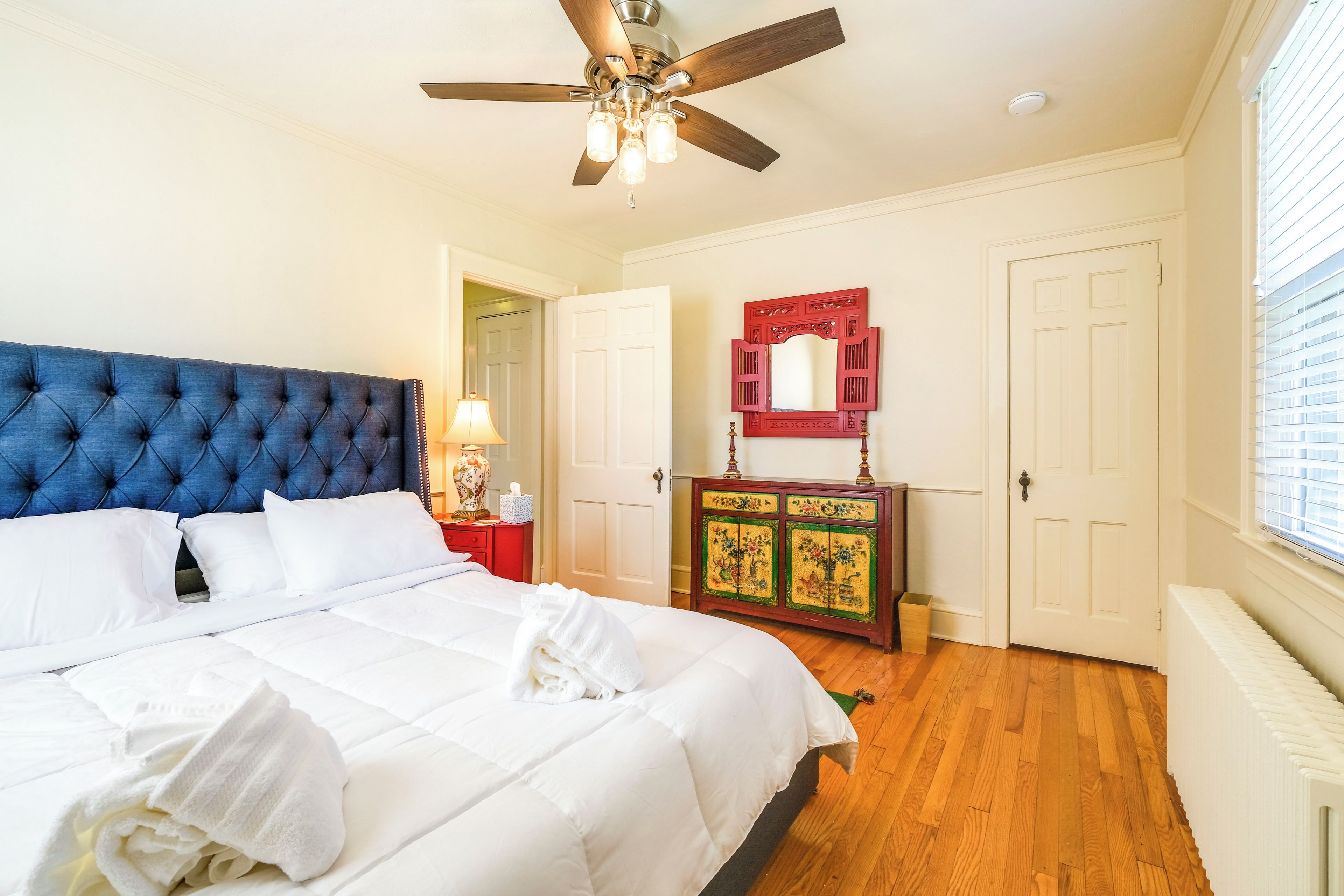 A bedroom with a tufted headboard, a ceiling fan, a wooden cabinet with a mirror, and a radiator.