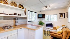 A modern kitchen with white cabinets, a wooden countertop, and a built-in stove. There is a TV mounted on the wall, a large window, and a brown sofa in the living area. - Hike On-site: Georgia Tiny Home on Farm Retreat (Lithonia)
