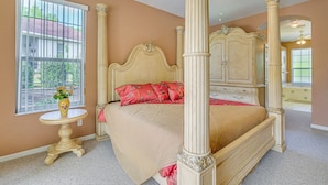 A four-poster bed with a beige bedspread and red pillows, a small round table with a vase, and a window with bars.
