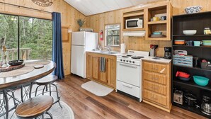 A cabin-style kitchen with wooden cabinets, a refrigerator, a stove, and a microwave. There is a dining area with a table and chairs, and a view of the forest through the window.