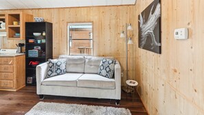 A cozy living room with a sofa, wooden walls, a dining area, and a kitchen with shelves and appliances.