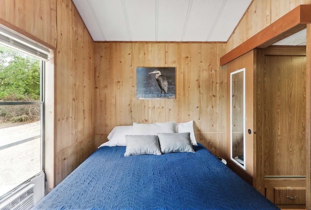 A bedroom with a wooden wall, a bed with blue bedding, and a picture of a bird on the wall.