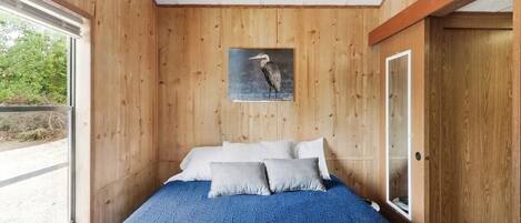 A bedroom with a wooden wall, a bed with blue bedding, and a picture of a bird on the wall.