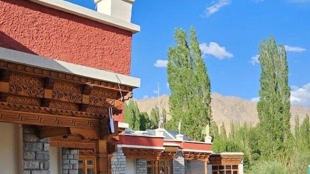 A traditional multi-story building with wooden balconies and stone walls, set against a backdrop of trees and mountains. - Hotel Leh Residency (Leh)