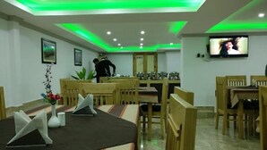 A dining area with wooden furniture, a TV mounted on the wall, and a person standing near the back. - Hotel Leh Residency (Leh)