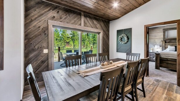 Un coin repas avec une table et des chaises en bois, une vue sur l’extérieur grâce à de grandes portes-fenêtres coulissantes, et un lit avec une tête de lit dans la pièce adjacente.