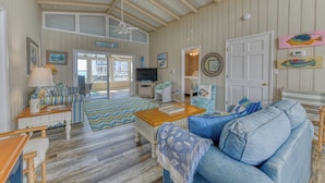 A spacious living room with a large sofa, a coffee table, and a TV. There is a door leading to a balcony, and the room has wooden floors and a high ceiling with a fan. - Carolina Palms - 5 Bedroom Second Row Beach House (Garden City Beach)