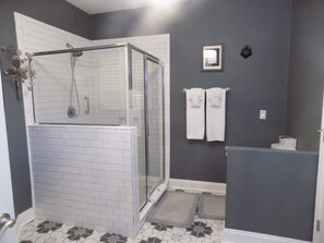 A bathroom with a glass-enclosed shower, white tiled walls, a gray accent wall, and a patterned floor. - The Lantern (Pensacola)