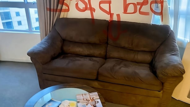 A brown sofa bed in a room with a window, a round glass table with books and tissues, and a wall with a partial view of a building outside.