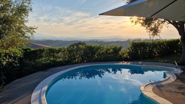 A curved swimming pool with a wooden deck, surrounded by greenery and a scenic view of distant hills. - Villa Gelso: your luxury villa with pool and sauna in Le Marche region (Provincia di)