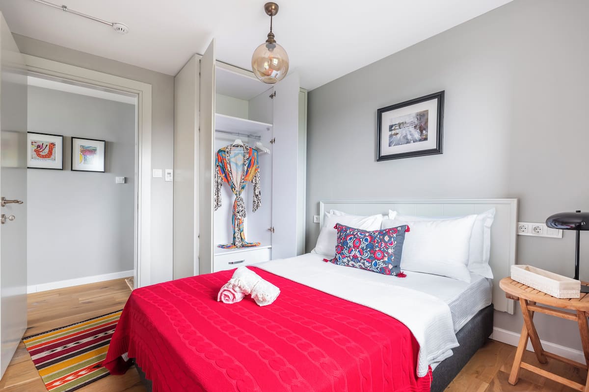 A bedroom with a bed, a red bedspread, a white pillow, a bedside table, and a hanging light fixture.