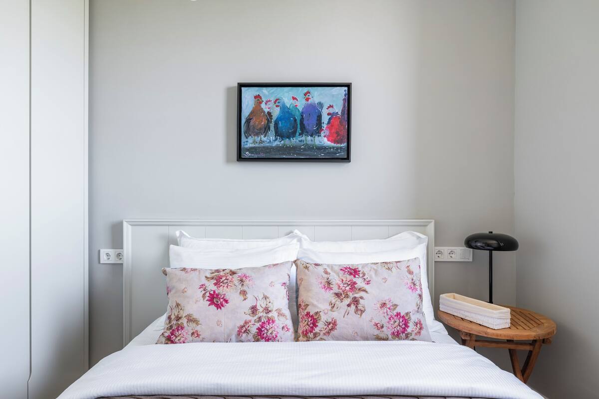 A neatly made bed with floral pillows, a framed artwork above the bed, and a small wooden side table with a lamp.