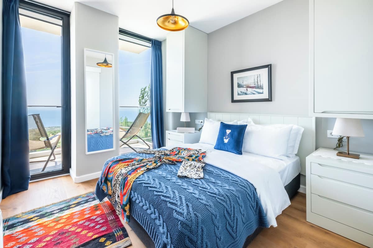 A modern hotel room with a large bed, a view of the sea, a balcony with a chair, and a painting on the wall.