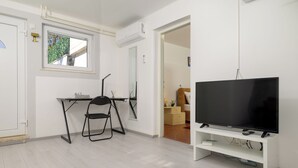 Ett modernt vardagsrum med en platt-TV på ett vitt TV-bord, en svart hopfällbar stol, ett litet skrivbord med en lampa och ett fönster med utsikt över grönområden.