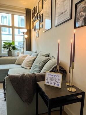 Ein gemütliches Wohnzimmer mit einer Couch, einem kleinen Tisch mit Kerzen und einem bordgestellten Schild mit der Aufschrift 'Willkommen'.