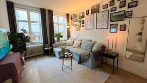 Een woonkamer met een bank, een salontafel, een potplant, een radiator, een raam met gordijnen en ingelijste foto's aan de muur.