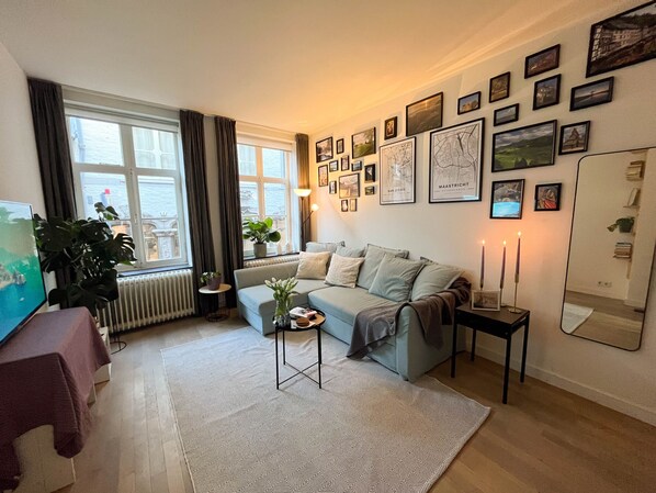 Ein Wohnzimmer mit einer Couch, einem Couchtisch, einem Topf mit Pflanze, einem Heizkörper, einem Fenster mit Vorhängen und gerahmten Bildern an der Wand.