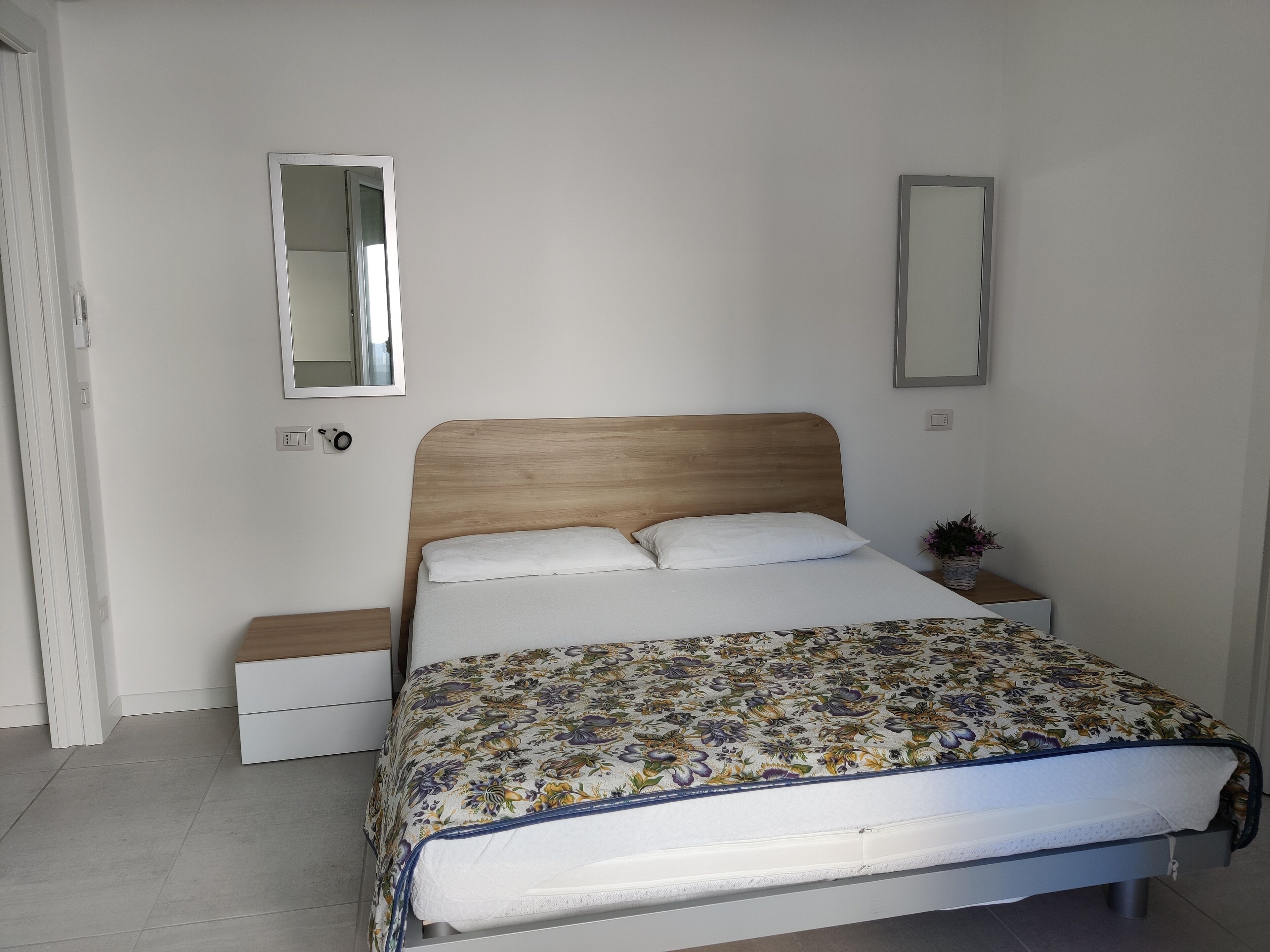 A neatly made bed with a floral bedspread, a wooden headboard, and a nightstand with a plant.