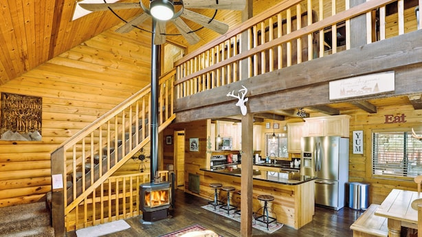 Interior de una cabaña de troncos con escalera de madera, chimenea, zona de cocina y comedor.