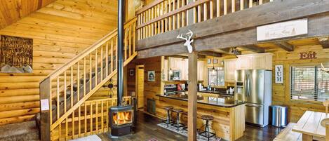 A log cabin interior with a wooden staircase, a fireplace, a kitchen area, and a dining space.