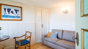 A room with a wooden floor, a grey sofa with a patterned cushion, a wooden chair with a blue cushion, a framed picture of a cow, and a wall-mounted light fixture. - Io&Te wellness space (Sordevolo)