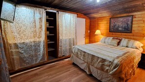 A wooden-paneled bedroom with a bed, two bedside lamps, a wardrobe with floral curtains, and a painting on the wall. - Holly's Hideaway, Cabin in the Woods (Nogal)