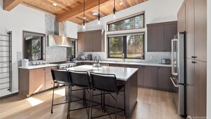 A modern kitchen with a large island, wooden ceiling beams, and a view of the outdoors. - 5 King Beds on Golf Course with a VIEW 0.6 mile walk to Winery! (Cle Elum)