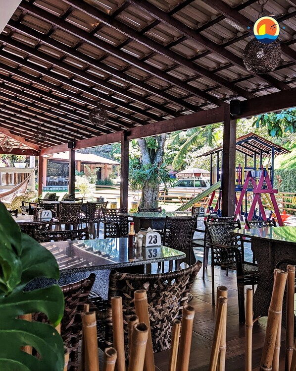 An outdoor restaurant with wooden tables and chairs, a covered area with a wooden ceiling, and a view of a playground in the background. - Pousada Ponta de Areia (Vila Velha)