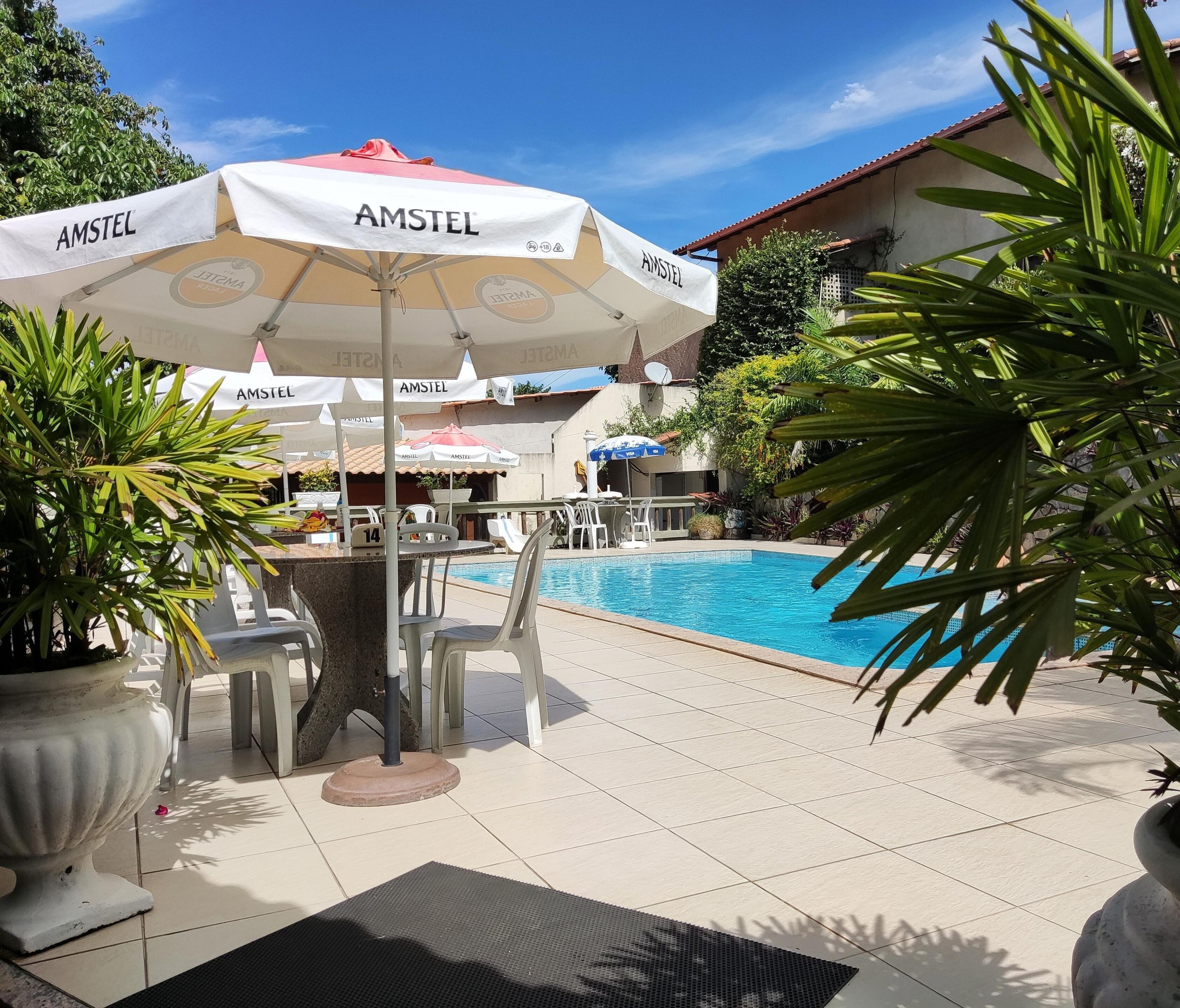 A poolside area with AMSTEL branded umbrellas, white plastic chairs, and a tiled floor.