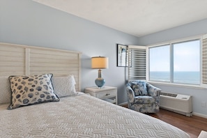 A bedroom with a bed, a nightstand, a chair, a window with blinds, and a view of the sea.