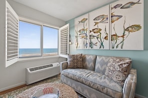 A living room with a view of the ocean, a white air conditioner, and a patterned sofa.